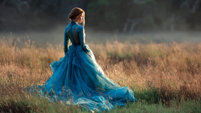 A bride in a long blue wedding dress walks through a sunlit field