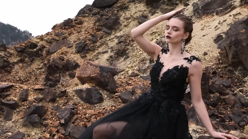 A bride in a black lace gown poses outdoors on rocky terrain, wearing a crown and statement earrings